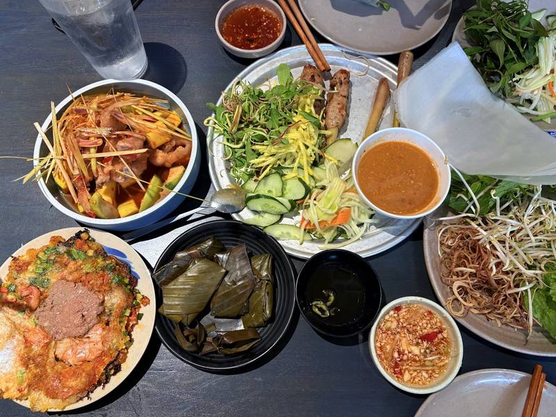 Bàn ăn đầy bánh ép, nem lụi, bánh lá, rau sống và nước chấm